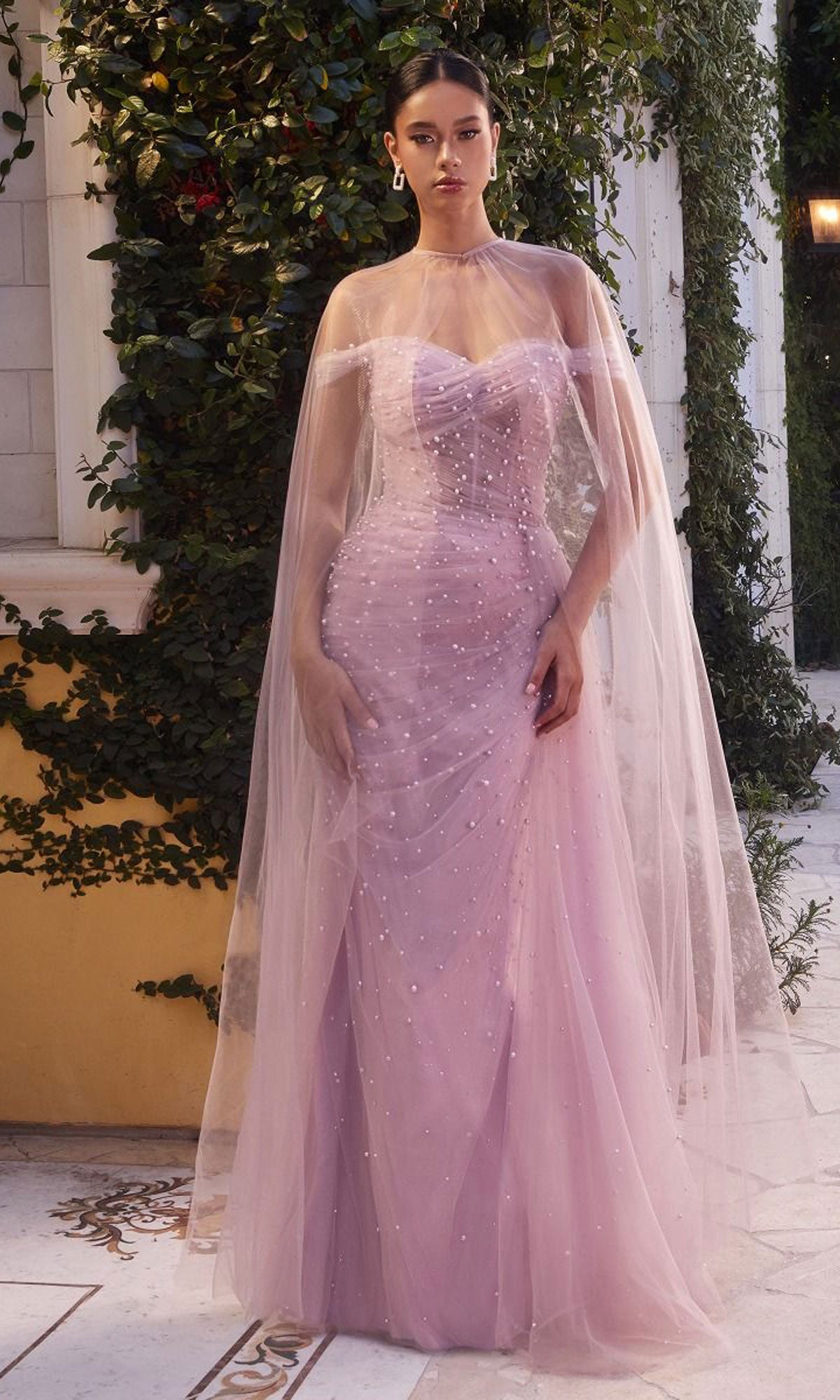 A woman stands outdoors in a Long Pink Pearlized Formal Dress with Cape, surrounded by greenery and architectural elements.